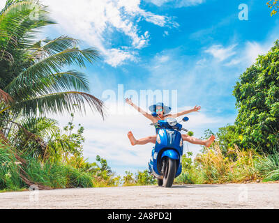 Crazy lustige Frau mit fliegenden Haare Motorrad fahren auf einem blauen Himmel und grüne Tropen Hintergrund. Junges Mädchen mit dunklen Haaren in Sonnenbrille auf einem blauen Stockfoto