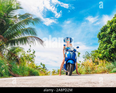 Crazy lustige Frau mit fliegenden Haare Motorrad fahren auf einem blauen Himmel und grüne Tropen Hintergrund. Junges Mädchen mit dunklen Haaren in Sonnenbrille auf einem blauen Stockfoto