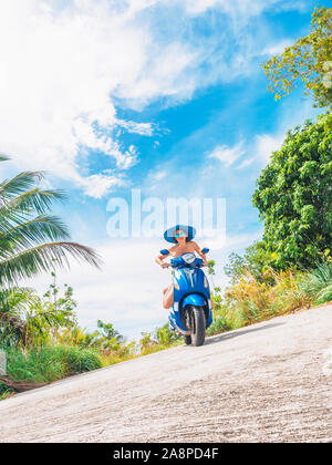 Crazy lustige Frau mit fliegenden Haare Motorrad fahren auf einem blauen Himmel und grüne Tropen Hintergrund. Junge bizarre Mädchen mit dunklen Haaren in Sonnenbrille auf ein Stockfoto