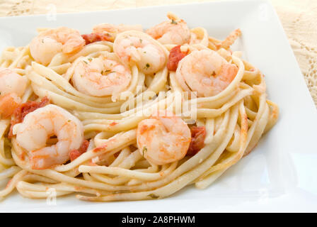 Hausgemachte Shrimp Scampi serviert auf einem Bett von Linguini pasta Nudeln in eine weiße quadratische Platte mit einer antiken Tischdecke im Hintergrund. Stockfoto