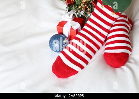 Weihnachtsfest zu Hause, überquerte weibliche Füße in rot-weiß gestreifte Socken und Neues Jahr Baum auf einem Bett. Frau in Santa Kleidung und festliche Dekoration Stockfoto