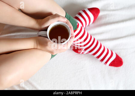 Tasse Tee in weibliche Hände, Frau mit heißen Getränk sitzen auf dem Bett cross-in Weihnachtssocken Beinen. Konzept der Silvester, zu Hause entspannen, gemütlich Stockfoto