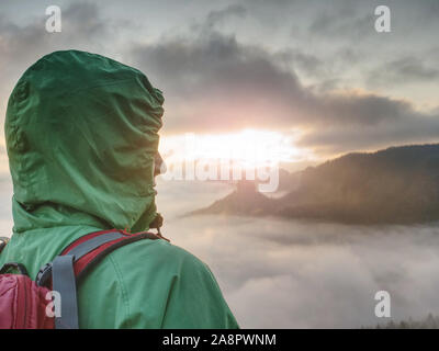 Freundin tragen grüne wasserdichte Jacke oder Regenjacke mit Kapuze und Rot mit Rucksack. Müde Wanderer Stop für kurze Pause auf felsigen Gipfel und genießen Stockfoto