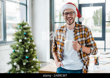 Selektiver Fokus der fröhlichen bärtigen Mann in santa hut Holding Cup in der Nähe von Weihnachten Baum im Büro Stockfoto