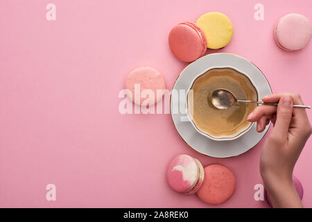 7/8-Ansicht von Frau mit silbernen Löffel über Kaffee Tasse in der Nähe von köstlichen französischen Makronen auf rosa Hintergrund Stockfoto