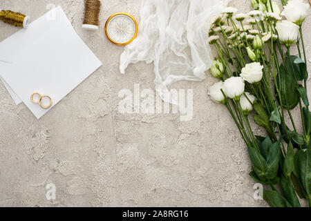 Blick von oben auf die Trauringe auf leere Einladungskarte, Weiß Gaze und Blumen auf strukturierte Oberfläche Stockfoto