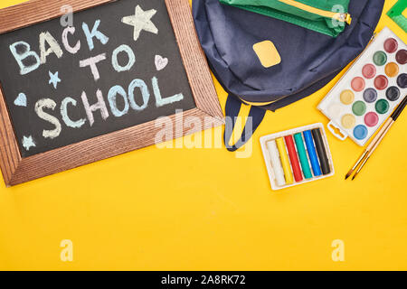 In holzrahmen Schiefertafel mit zurück in die Schule Inschrift in der Nähe von Blue Schulranzen und Aquarellfarben auf gelbem Hintergrund Stockfoto