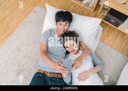 Blick von oben auf die Glücklichen asiatischen Liebhaber verwenden die Technologie Smart Phone für selfie auf dem Fußboden mit Teppich im Schlafzimmer zu Hause, Paar selfie Konzept, Stockfoto