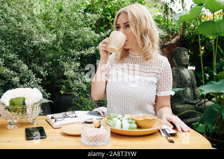 Schöne elegante junge Frau trinkt Kaffee und Essen Frühlingsrollen für Business Lunch im Café im Freien Stockfoto