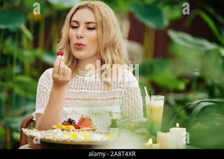 Reizende junge blonde Frau am Tisch sitzen im Café draußen und Blasen auf Erdbeere, bevor es Essen Stockfoto