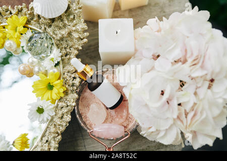 Flasche mit feuchtigkeitsspendenden Essenz in der Schüssel von rose Pulver und Massage Roll auf Tisch mit frischen Blumen, Ansicht von oben Stockfoto