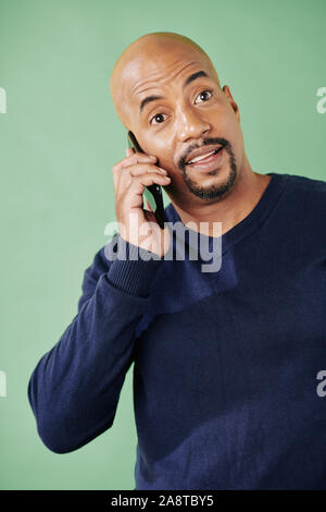 Portrait von positiven kahlen Mixed-race Mann am Telefon sprechen mit Freund oder Verwandten, isoliert auf Grün Stockfoto