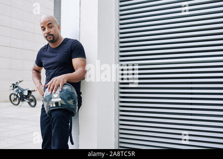 Stattlicher Mann mit Glatze Rundum-bart snatding mit Motorrad Helm in der Hand und Suchen an Kamera Stockfoto