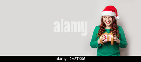 Glücklich lächelnde Frau in Rot Weihnachtsmütze und grünen Pullover hält Weihnachten Geschenk Box mit Gold Band auf einem grauen Hintergrund mit kopieren. Xmas holiday Co Stockfoto
