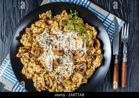 Steinpilzen cremige Risotto mit geriebenem Käse und Thymian auf schwarzem Teller auf einem Holztisch, Ansicht von oben, flatlay, close-up Stockfoto