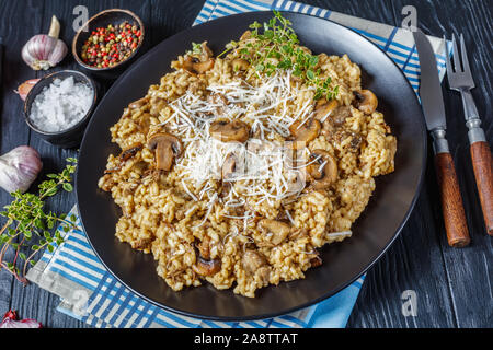 Steinpilzen cremige Risotto mit geriebenem Käse und Thymian auf eine schwarze Platte mit Zutaten auf einem Holztisch, Ansicht von oben, close-up Stockfoto