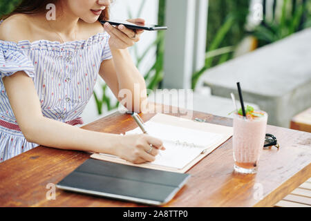 Zugeschnittenes Bild der weiblichen Studenten im Cafe sitzen, Aufzeichnung von Sprachnachrichten und Schreiben in Planer Stockfoto