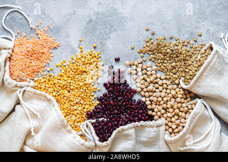 Verschiedenen Hülsenfrüchten in eco Beutel. Linsen, Bohnen, Erbsen, Kichererbsen sind auf einem grauen Hintergrund verstreut. Flach mit Platz für Text. Stockfoto