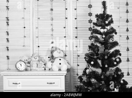 Feiern und das Neue Jahr Einrichtung Konzept. Schneemänner und Teddybären in der Nähe der Uhr und Weihnachtsbaum. Spielzeug auf Präsidium auf Holz Wand und Girlanden Hintergrund. Tannenbaum mit Dekorationen im festlichen Zimmer Stockfoto