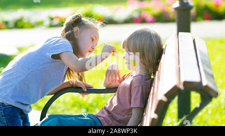 Ein kleines Mädchen ist die Anwendung aqua Make-up im Gesicht von einem 5 Jahre alten Jungen, der auf einer Bank sitzen ein Park ist. Stockfoto