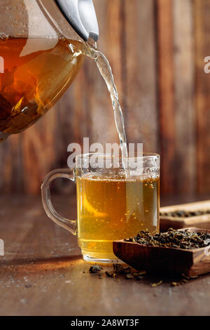 Frischer grüner Tee wird aus einer Teekanne, die in einem Becher gegossen. Hot grüner Tee im Glas Tasse auf einem Holztisch. Stockfoto