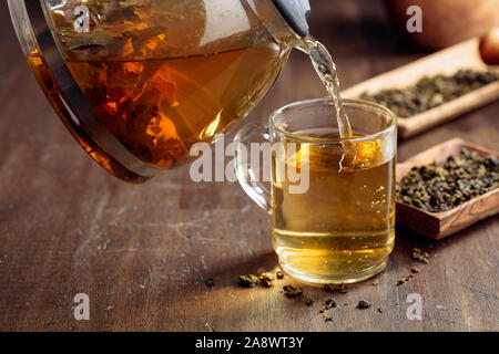 Frischer grüner Tee wird aus einer Teekanne, die in einem Becher gegossen. Hot grüner Tee im Glas Tasse auf einem Holztisch. Stockfoto