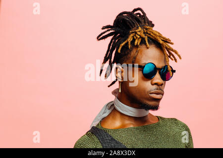 Nahaufnahme, Porträt eines jungen Mannes in Grün mit Dreadlocks und blauer Sonnenbrille, isoliert auf Rosa Stockfoto