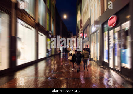 Blurry motion Image der jungen Menschen zu Fuß auf der Kalverstraat Straße, einer der wichtigsten Einkaufsstraßen in Amsterdam. Es ist ein regnerischer Sommer Nacht. Stockfoto