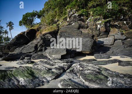 Hanglage mit schwarzen Felsen und grünen Bäumen, Pflanzen am Ufer des wunderschönen exotischen Sandstrand und eine atemberaubende Cenang Beach in Insel Langkawi, in Malaysi Stockfoto