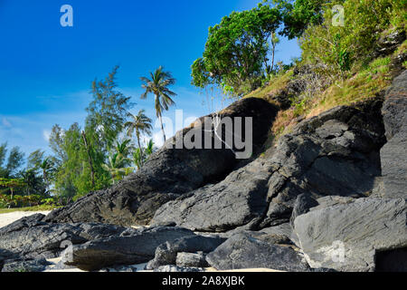Hanglage mit schwarzen Felsen und grünen Bäumen, Pflanzen am Ufer des wunderschönen exotischen Sandstrand und eine atemberaubende Cenang Beach in Insel Langkawi, in Malaysi Stockfoto