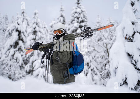 Foto von Athleten in Helm und Maske mit Skiern auf seiner Schulter gegen den Hintergrund der Bäume im Schnee im Winter Resort Stockfoto