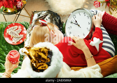 Obere Ansicht von Mutter mit Kaffee Tasse streicheln Tochter, die schlafend Warten auf Weihnachten fiel beim Sitzen auf einem Sofa in der modernen Wohnzimmer in Christus Stockfoto