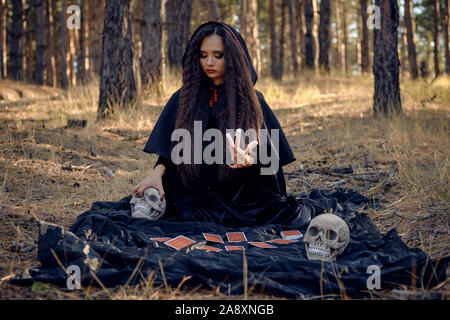 Attraktive, böse, langhaarigen Charmeur in einem schwarzen, langen kleid mit umhang und Kapuze. Sie hält einige Kerzen, sitzen auf einer dunklen Decke whith Fortun Stockfoto