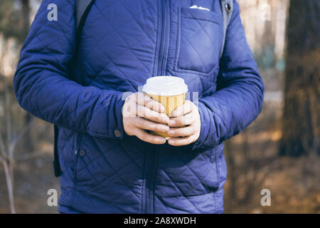 Die menschliche Hand hält ein Papier Kaffee Tasse mit Kunststoffkappe, Social Media. Pappbecher in die Hände der Mann in der blauen Jacke. Getönten Bild. Stockfoto