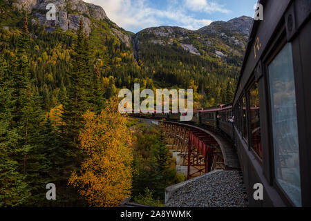 Skagway, Alaska, Vereinigte Staaten - 24 September 2019: Alte historische Eisenbahn Zug fährt über eine Holzbrücke bis White Pass mit Touristen während einer cl Stockfoto