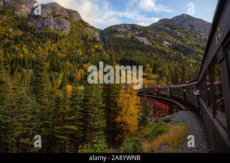 Skagway, Alaska, Vereinigte Staaten - 24 September 2019: Alte historische Eisenbahn Zug fährt über eine Holzbrücke bis White Pass mit Touristen während einer cl Stockfoto