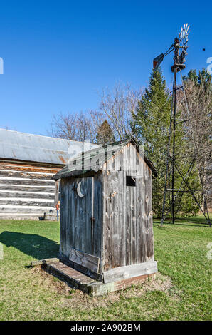 Verwitterte Plumpsklo mit einem weißen Halbmond auf der Tür und eine Windmühle im Hintergrund Stockfoto