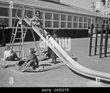 Kinder spielen im Freien. Eine afrikanische amerikanische Kind ist Rutschen eine Folie während andere Kinder auf Matten rund um die Folie werden kann. 1950 s Stockfoto