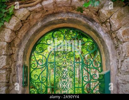 Street View eines dekorativen Iron Gate in einem rustikalen, gewölbte Stein Eingang zu einem Sommer Garten mit üppigem Grün im hellen Licht erfüllte, in Italien. Stockfoto