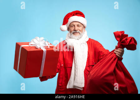 Gerne reife Santa Claus in Red Hat, Jacke und weißen Strickschal Hände halten in roter Geschenkbox und der große Sack, auf blauem Hintergrund isoliert Bärtigen. Urlaub präsentiert und Weihnachten. Stockfoto