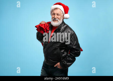 Zuversichtlich reife bärtige Mann in rote Mütze, Lederjacke und Jeans an Kamera suchen und halten grosse Santa Sack voller Geschenke. Zeit der Happy winter Urlaub und schöne Geschenke Stockfoto