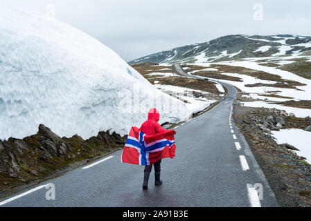 Bjorgavegen - verschneite Straße in Norwegen. Mädchen mit die norwegische Flagge auf einem Berg Straße in der Nähe einer Wand aus Schnee. Schwere nördlichen Landschaft Stockfoto