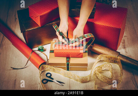 Nahaufnahme der Frau Verpackung Weihnachtsgeschenke in Papier. Menge Kartons, Bänder, Schere, Klebeband auf weißem Holzfußboden. Stockfoto