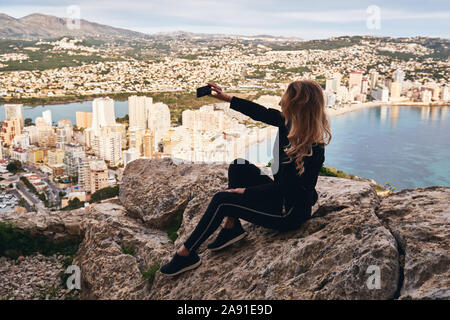 Blonde Frau auf Stein rock Posing macht selfie Bild Mobiltelefon erfassen Moment kletterte auf Penon de Ifach Rock im Park sitzen. Calpe spanis Stockfoto