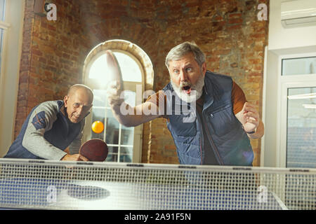 Ältere Männer Tischtennis spielen am Arbeitsplatz, Spaß zu haben. Freunde in legere Kleidung spielen Ping pong zusammen an einem sonnigen Tag. Konzept der Freizeitgestaltung, Sport, Freundschaft, Teambuilding, Teamarbeit. Stockfoto
