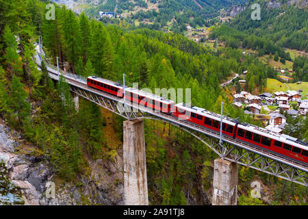 Zermatt, Schweiz - Oktober 7, 2019: Gornergrat rote Bummelzug auf der Brücke, Häuser des Dorfes und Schweizer Alpen Panorama Stockfoto