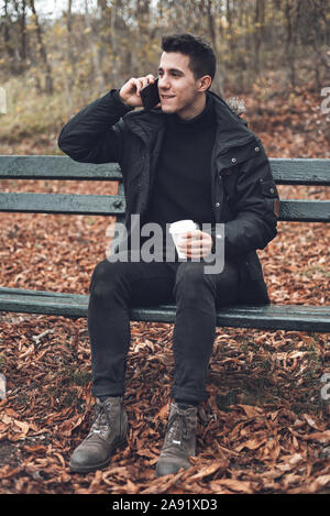 Junge lächelnde Mann mit Mantel mit Handy und trinken Kaffee zum Mitnehmen im Herbst Park. Stockfoto