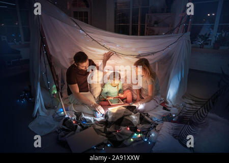 Kaukasische Familie sitzt in einem Tipi, Spaß und Spielen mit der Taschenlampe im dunklen Zimmer mit Spielzeug und Kissen. Glücklich aus. Home Komfort, Familie, Liebe, Weihnachten, Storytelling. Stockfoto