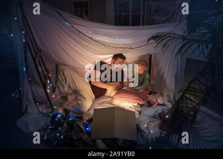 Vater und Tochter sitzen in einem Tipi, Spaß haben, spielend mit der Taschenlampe im dunklen Zimmer mit Spielzeug und Kissen. Glücklich aus. Home Komfort, Familie, Liebe, Weihnachten, Storytelling. Stockfoto