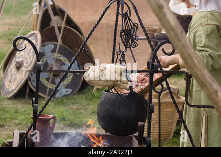 Frau gekleidet in mittelalterlichen Kostüm Vorbereiten der Nahrung zu einem Re-enactment Camp mit einem Spieß gebratenes Huhn und Kessel Stockfoto